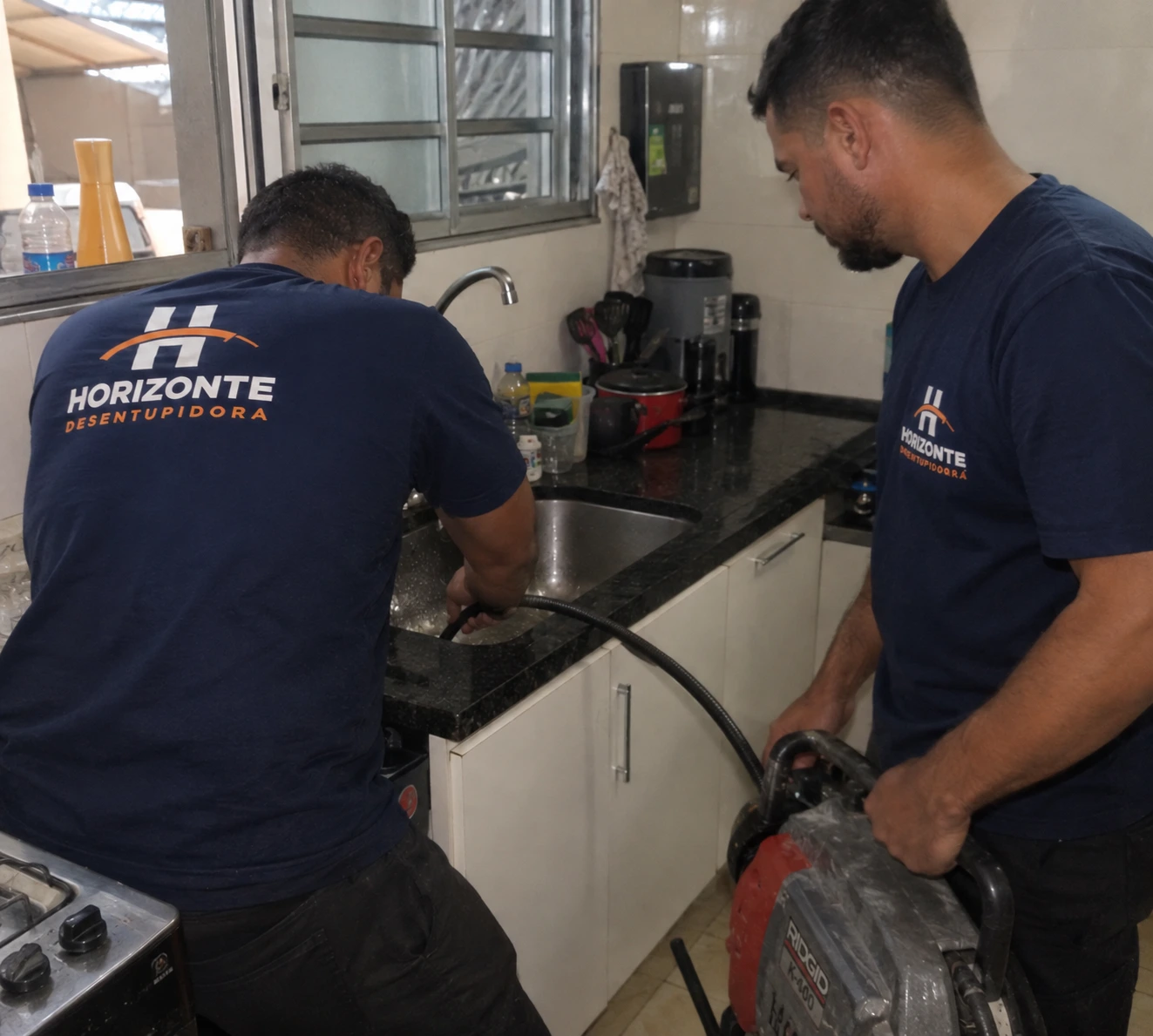 Dois técnicos da Horizonte desentupindo a pia de uma cozinha com máquina rotativa.